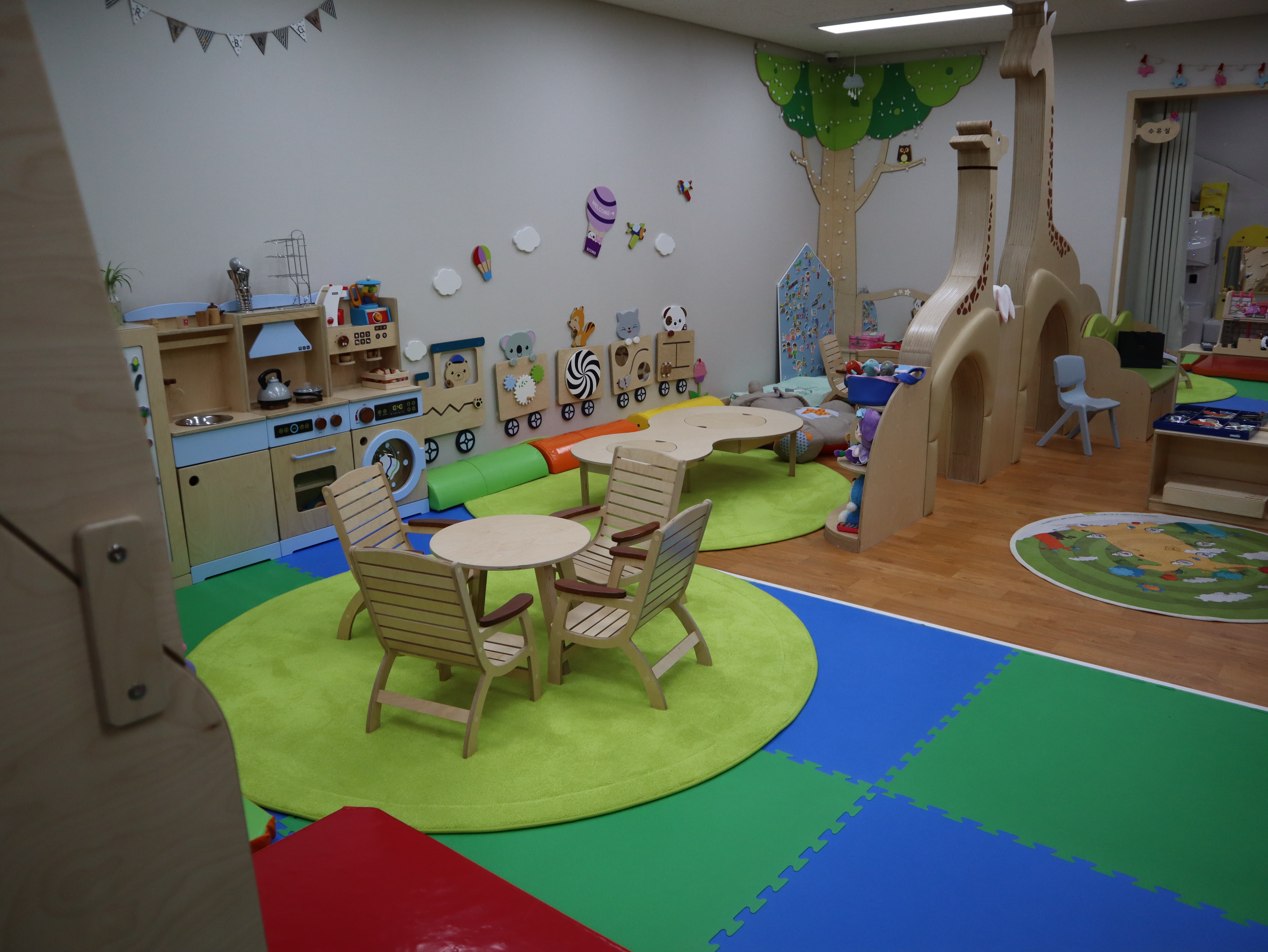 유아놀이공간0 : 폭신한 바닥재가 깔려 있고 중앙에 원형 테이블과 의자가 있는 유아 놀이방 내부