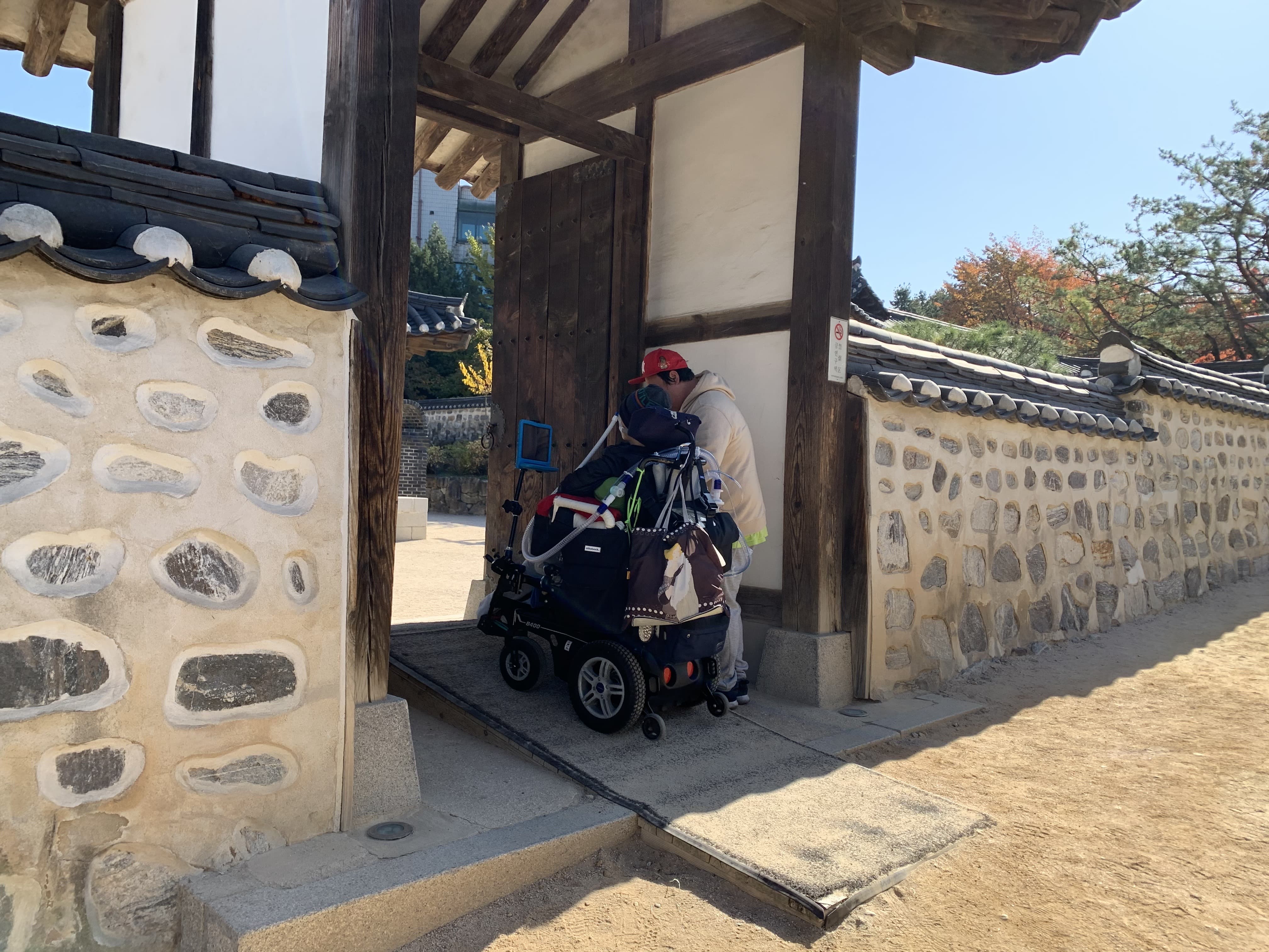 이동로0 : 전통가옥 출입구에 설치된 짧은 길이의 경사로를 지나가는 휠체어 사용자와 동반자