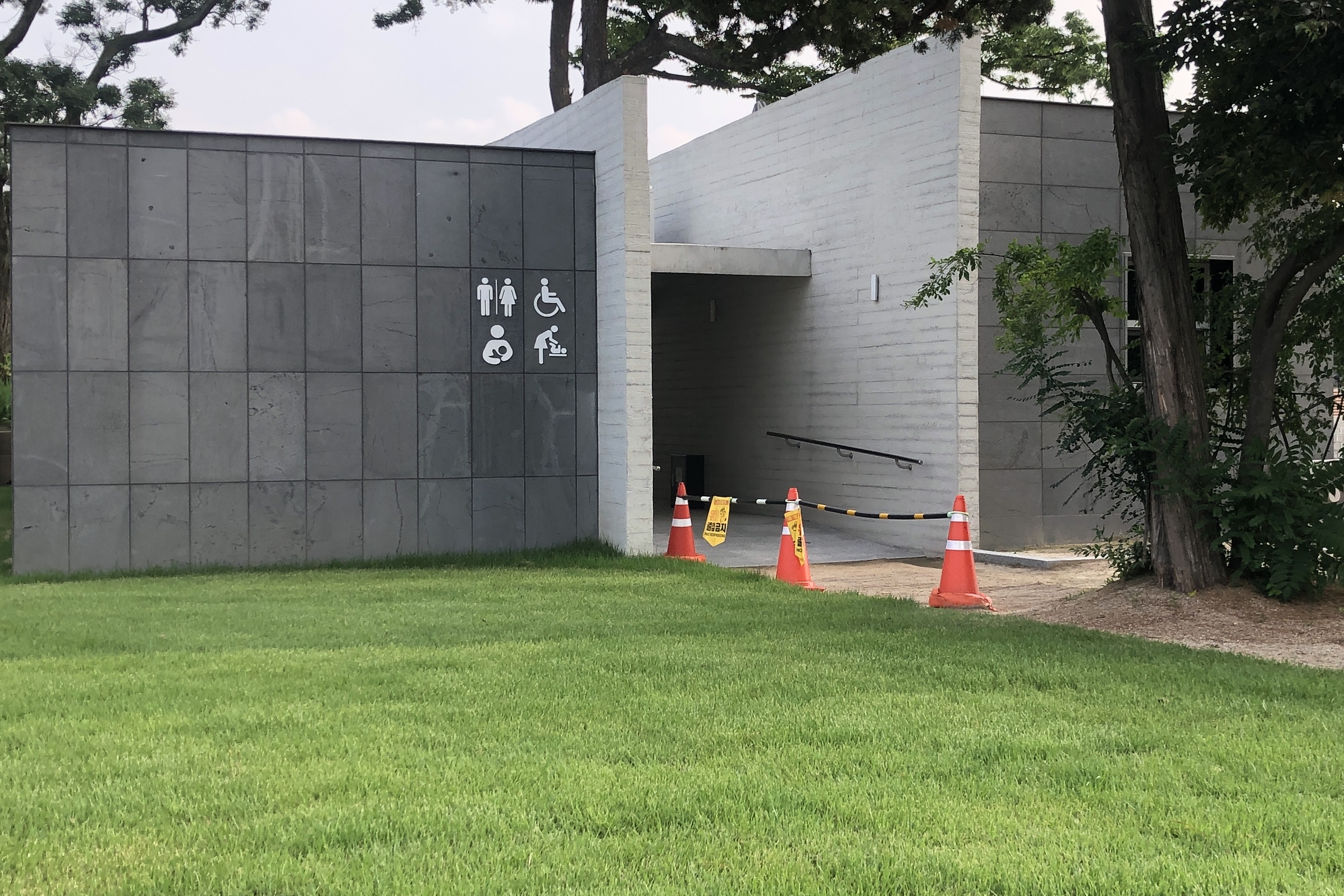 Accessible restroom for persons with disabilities0 : Toilet building in front of lawn with pictograms of toilet, wheelchair, nursing room, and diaper changing station

